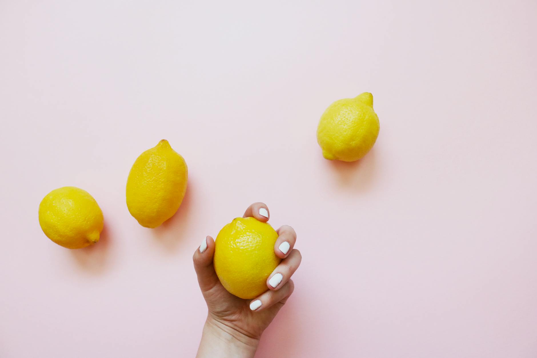 Hand holding a fresh lemon on pink background with additional lemons nearby, symbolizing natural pleasure and cycles