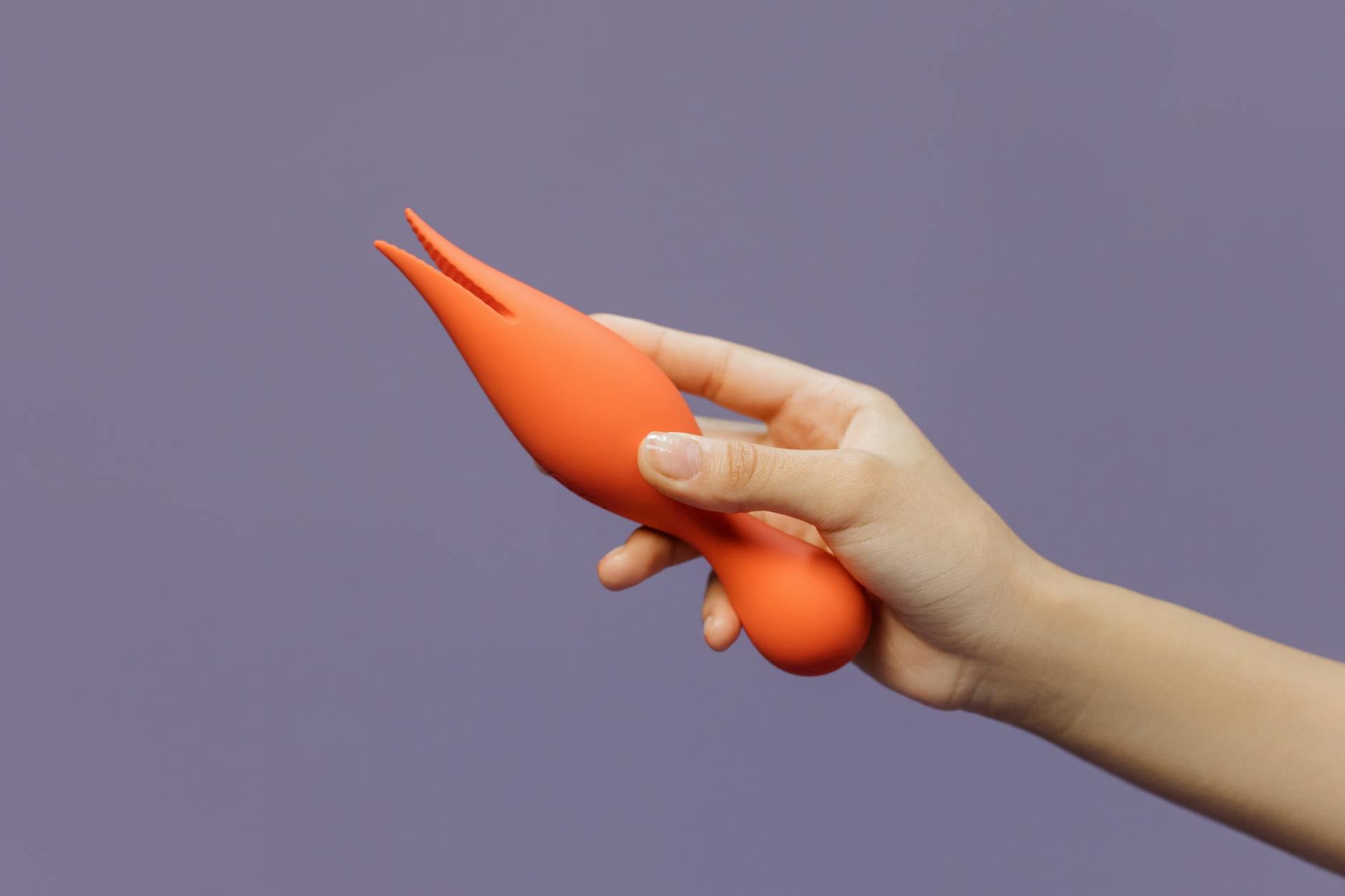 Close-up of a hand holding an orange vibrator against a minimalistic purple backdrop, symbolizing solo pleasure and self-connection.