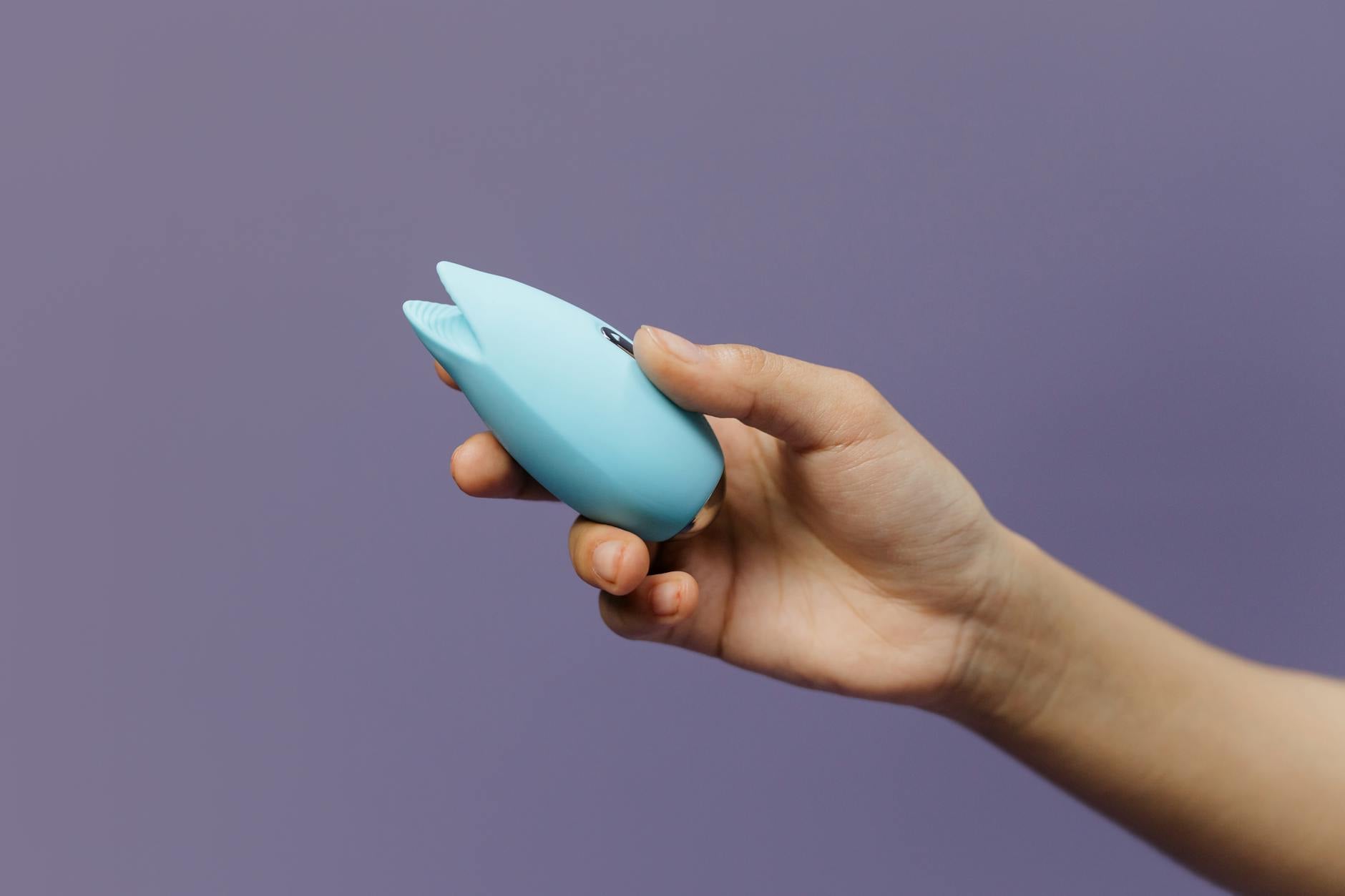 A silicone clitoral vibrator held in hand against a purple background, representing solo pleasure exploration