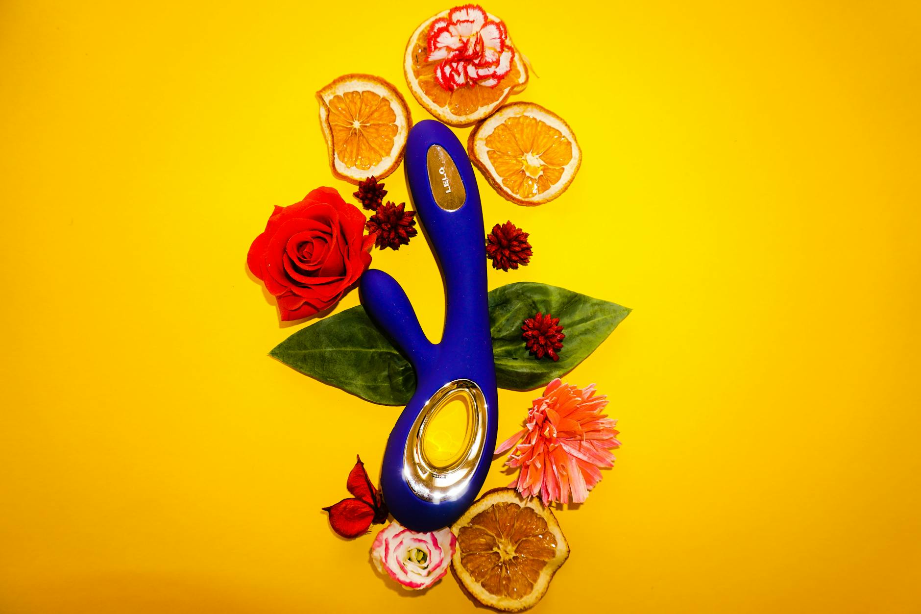 A person holding a blue vibrator surrounded by flowers and fruit slices on a yellow background